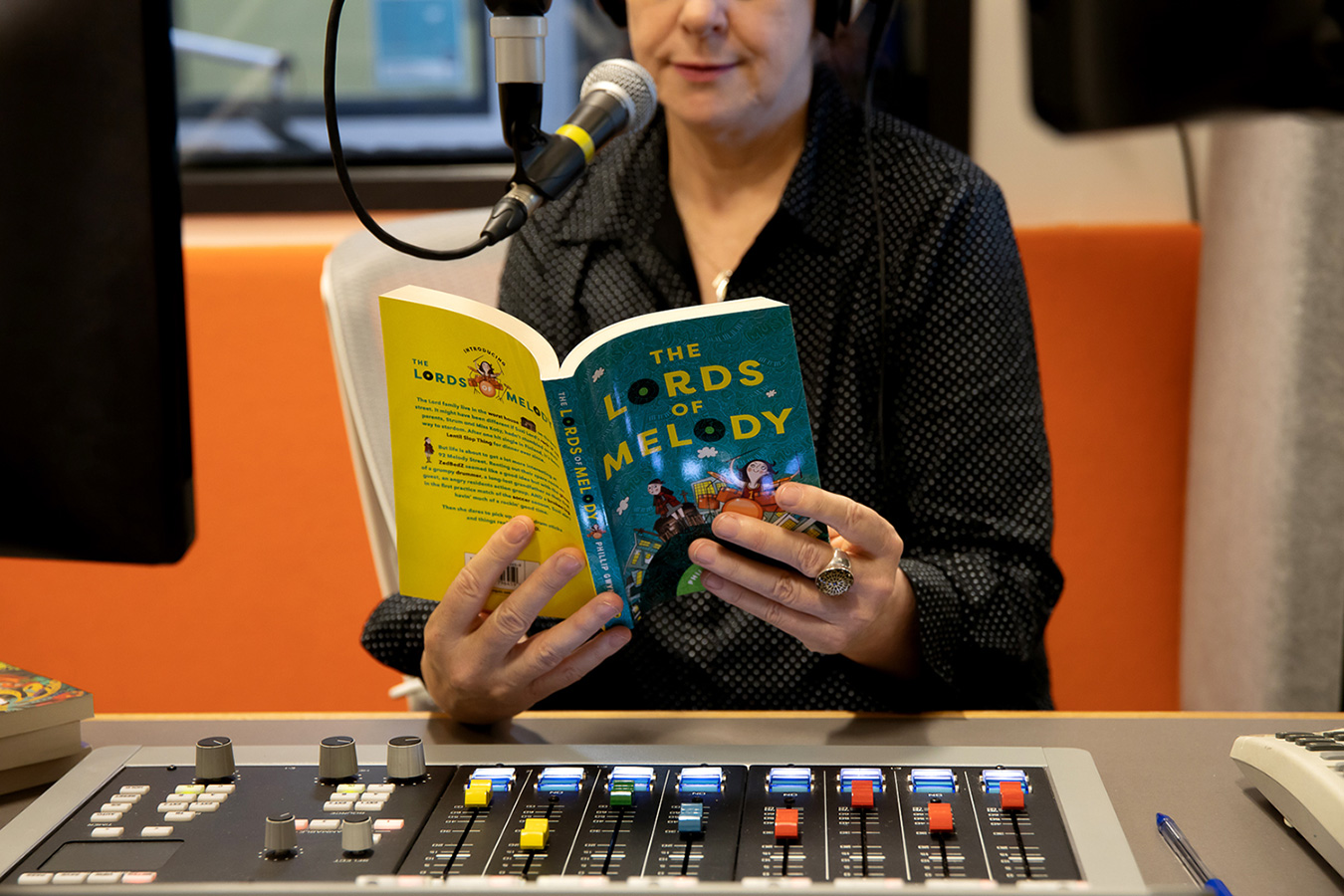 A woman in a black top sits at a desk with a microphone and an audio console in front of her. She is reading 'The Lords of Melody' by Phillip Gwynne