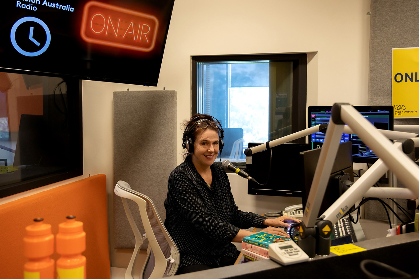 A middle-aged woman with dark hair and wearing a black top is smiling at the camera and sits at a radio studio desk. She is wearing headphones and a pile of books sits on the desk.