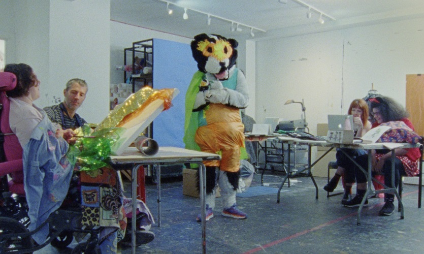 A still from the film The Stimming Pool, 2024, UK. A person in a handcrafted animal costume is standing in the middle of a a room. There are adults in the room who look to be making papier mache craft items