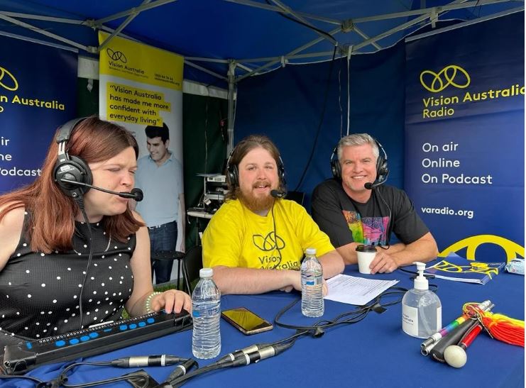 Three Vision Australia Radio team members sit together at a booth in a blue marquee smile at the camera.