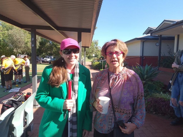 Vision Australia Radio volunteers Emma and Elizabeth smile at the camera while standing next to the lawn bowls bowling green.