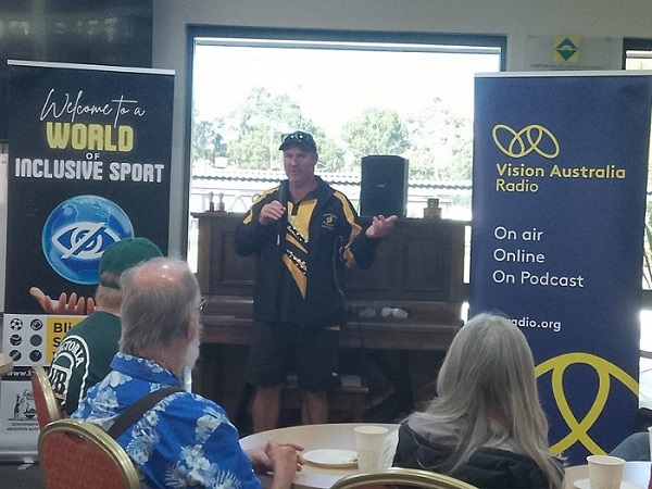 A man stands at the front of a room speaking to a number of volunteers in attendance. To his left is a Vision Australia pull up banner, and to his right is a Blind Sports pull up banner.