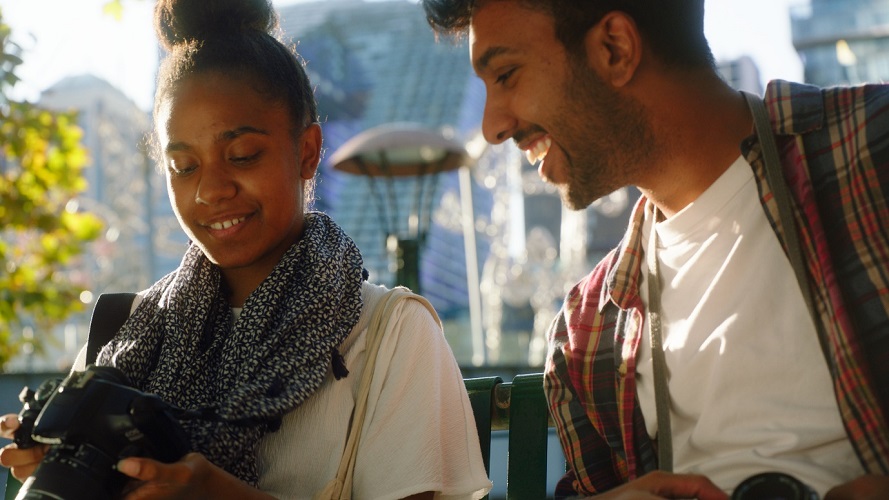 A still from the film Bokeh, 2024 Australia. A young woman is holding onto a camera and looking at the back screen. a Young man is standing next to her, and is also looking at the back of the camera, which the young woman has angled towards him.