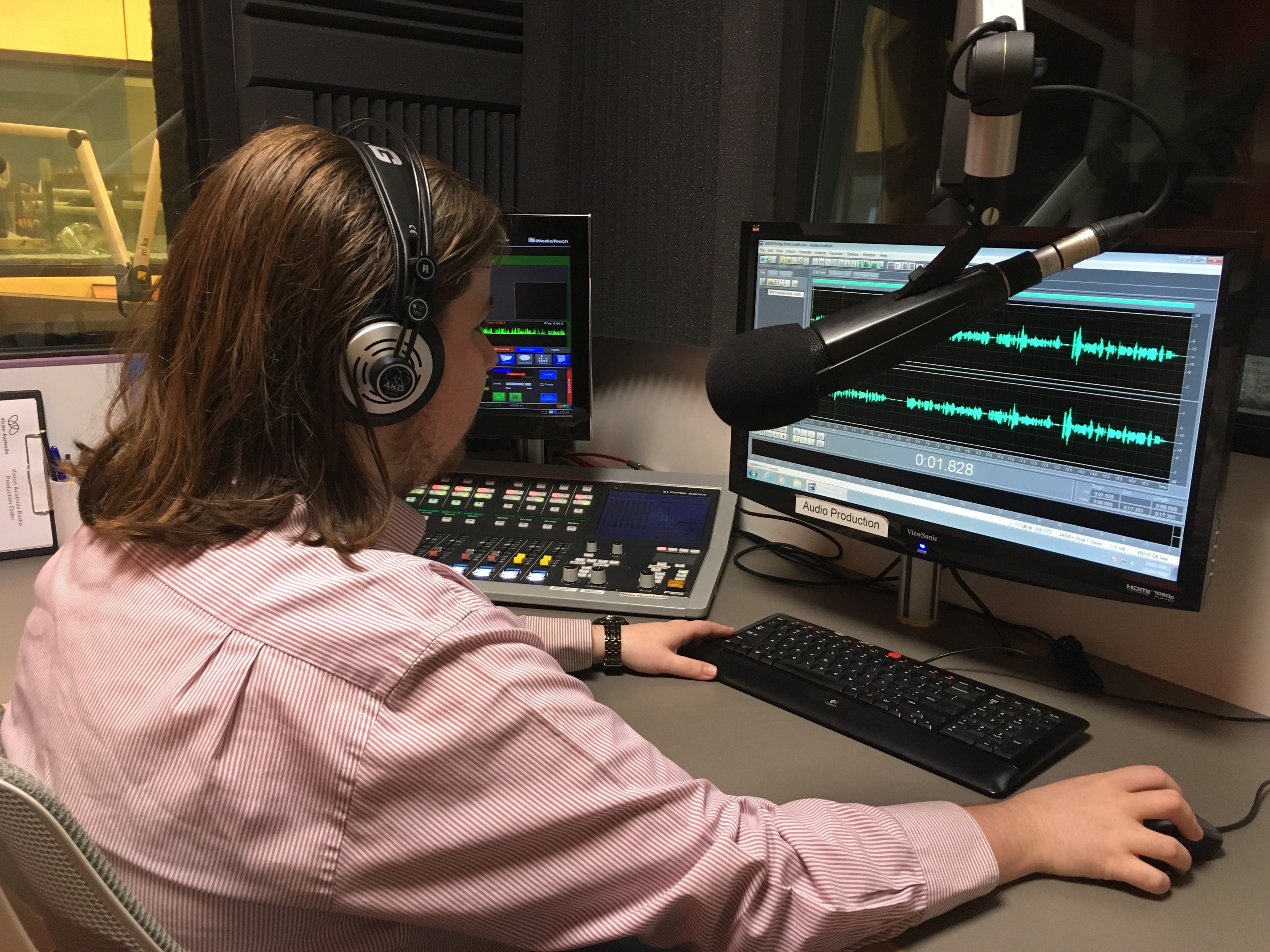 Vision Australia Radio graduate Sam Colley in the studio with headphones on, working on mixing desk and computer