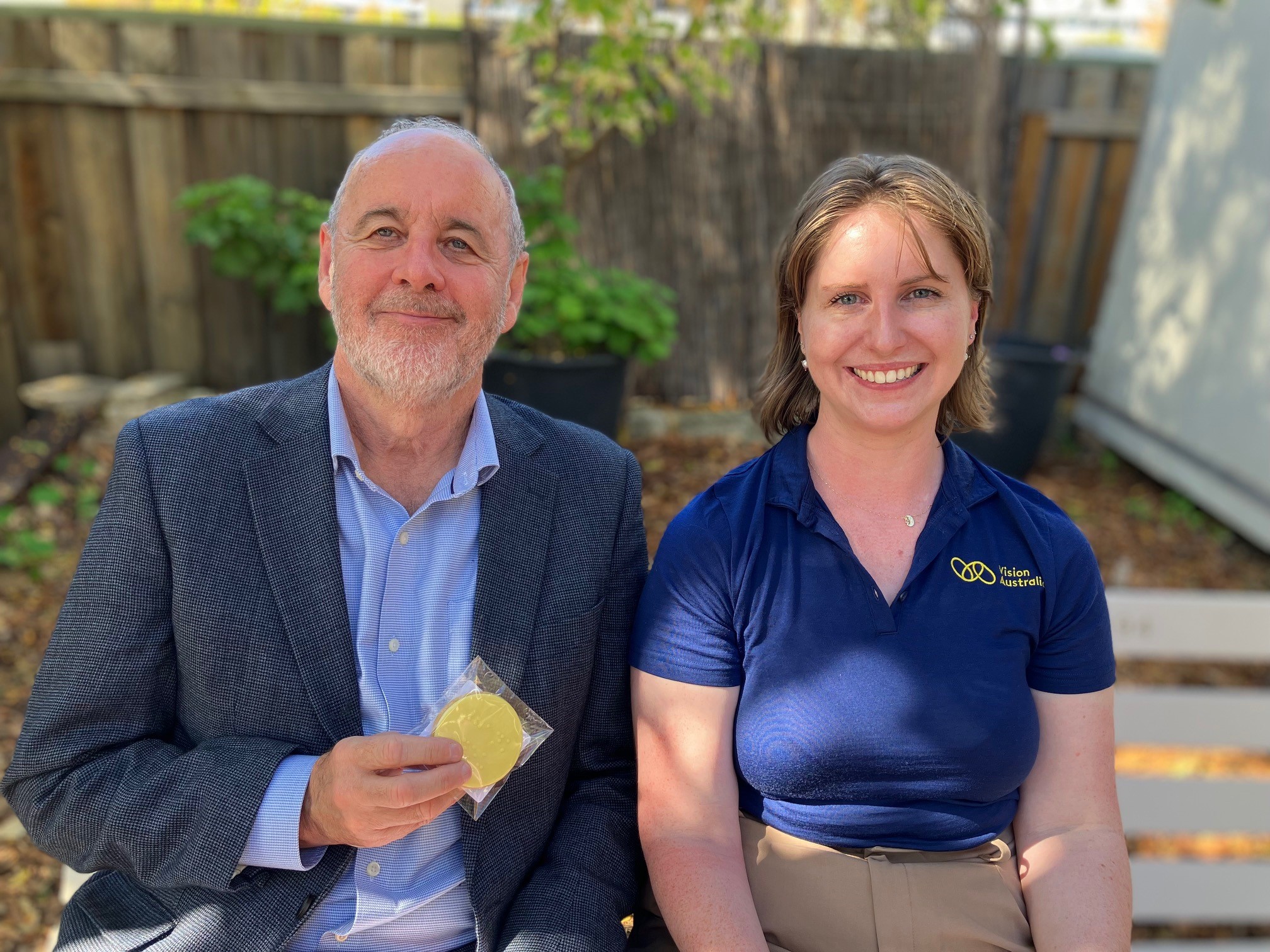 CEO Ron Hooton and Regional Client Services Manager Cara Patterson sit side by side. Ron is wearing a dark blue suit with blue top, Cara has shoulder-length brown hair and is wearing a blue Vision Australia polo t-shirt